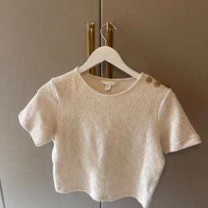 Vit croppad topp från H&M med knappar - Säljer en vit croppad topp från H&M med korta ärmar och rund hals. Toppen har en unik textur i frottéliknande material och tre dekorativa guldfärgade knappar på ena axeln. Perfekt för dig som gillar stilrena detaljer och en modern look.