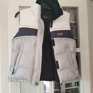 Peak Performance dunväst i grått och vitt - Snygg dunväst från Peak Performance i storlek L. Västen är quiltad och har färgblock i vitt, ljusgrått och mörkgrått med en röd detalj. Utrustad med dragkedja och hög krage, perfekt för kyliga dagar. Logga på bröstet och cool Peak Performance-zipper. Säljer den efter som den inte passar