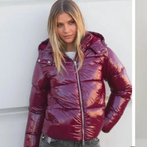 Glansig vinröd pufferjacka med huva - Säljer en glansig vinröd pufferjacka med huva och dragkedja framtill. Jackan har en kortare modell och breda kanaler, perfekt för kalla dagar. Snygg och trendig look som sticker ut i mängden.