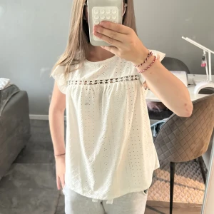 Vit blus med spetsdetalj - Säljer en söt vit blus med broderade hålmönster och en spetsrad med små blommor över bröstet. Blusen har korta ärmar med lätt volang och är tillverkad i mjuk bomull 💗