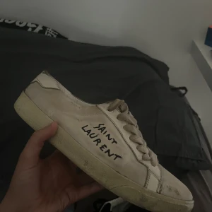 Saint Laurent vita sneakers med text - Säljer ett par vita sneakers från Saint Laurent med handskriven logga på sidan. Skorna har snörning, platt sula och är tillverkade i canvas med detaljer i skinn. Klassisk och avslappnad modell med rund tå och slitna kanter för en cool look.