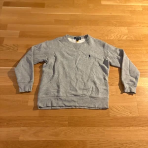 Grå sweatshirt från Polo Ralph Lauren - Klassisk grå sweatshirt från Polo Ralph Lauren med rund hals och broderad marinblå logga på bröstet. Tröjan har långa ärmar, ribbade muddar och är tillverkad i mjuk bomullsmix. Perfekt för en chill och stilren look.
