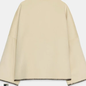 Beige kappa med svarta knappar - Stilren beige kappa med oversized passform och svarta knappar längs sidan. Jackan har markerade svarta sömmar längs kanterna och hög krage. Tillverkad i ett mjukt ullmaterial som ger en lyxig känsla. Perfekt för dig som gillar minimalistisk och modern stil.