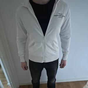 Vit hoodie från Peak Performance M - Vit hoodie från Peak Performance med dragkedja och huva. Klassisk logga på bröstet, två fickor framtill och ribbade muddar. Mjuk insida i bomullsmix, perfekt för chill dagar eller när du vill ha en clean look. Storlek M men den har krympt så den passar nog mer som storlek S. Liten fläck längst ner på framsidan, men inget som man tänker på. Hör av dig vid funderingar.