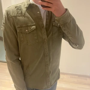Grön overshirt med slitningar - Säljer en svinfet grön overshirt som är omöjlig att få tag på i ett topp skick, med feta slitningar på axlar, ärmar och fickor. Skjortan har två bröstfickor med lock och tryckknappar samt klassisk krage. Denna är perfekt inför hösten