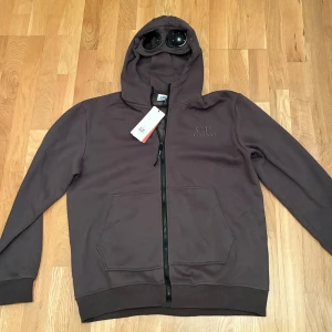 Brun hoodie med goggles från C.P. Company - Säljer en brun hoodie från C.P. Company med ikoniska goggles i huvan. Tröjan har dragkedja framtill, två stora fickor och broderad logga på bröstet. Tillverkad i mjuk bomull och perfekt för dig som vill sticka ut med streetwear-vibe.