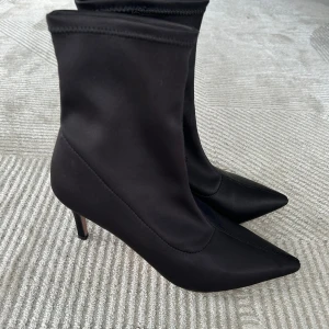 Svarta stövletter med spetsig tå - Snygga svarta stövletter med smal klack och spetsig tå. Skorna är i ett slätt, glansigt syntetmaterial som ger en elegant look. Modellen är åtsittande runt vristen och har en klassisk siluett som passar till många olika outfits.