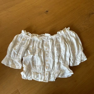 Vit offshoulder blus med knappar - Säljer en söt vit blus med offshoulder-modell och trekvartsärmar. Blusen har knappar framtill, smockdetaljer vid midja och ärmslut samt volangkant upptill. Använd en gång 