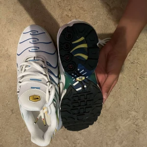 Nike Air Max Plus vita/blå/gula - Nike Air Max Plus sneakers i vitt mesh med blå och gröna vågformade detaljer samt gula accenter. Snygg synlig Air Max-sula och svart yttersula. Klassisk Nike-logga på sidan och tungan. Skorna har snörning och en sportig vibe, perfekt för dig som gillar streetwear.