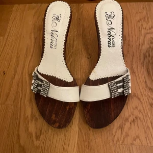 Vita sandaler med klack från Nelvas - Snygga sandaler från Nelvas med vit rem i skinn och glittriga strassdetaljer framtill. Sulan är i mörkt trä och klacken är smal i metall med coola nitar. Perfekt för dig som vill sticka ut med en unik och trendig look.