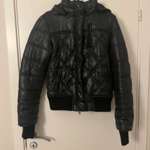 Svart quiltad pufferjacka  - Snygg svart pufferjacka med quiltat mönster och huva. Jackan har dragkedja framtill, ribbade muddar och detaljerade sömmar. Perfekt för kyliga dagar och ger en cool vibe till din outfit.