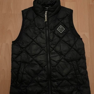 Svart quiltad dunväst från Gant - Svart quiltad dunväst från Gant med hög krage och dragkedja framtill. Västen har ett stilrent diamantmönster och en snygg Gant-logga på bröstet. Perfekt för lager-på-lager och passar till många olika stilar.