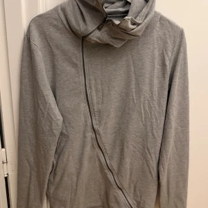 Grå tröja med sned dragkedja Army of Me - Säljer en grå hoodie från Army of Me i storlek M. Tröjan har en unik sned dragkedja framtill och är tillverkad i mjuk bomullsmix. Perfekt för dig som gillar stilrena och moderna detaljer. Långärmad och passar till lager-på-lager.