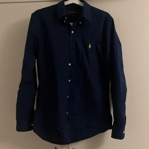 Mörkblå skjorta från Ralph Lauren - Säljer en mörkblå skjorta från Ralph Lauren i slim fit-modell. Skjortan har klassisk krage, vita knappar och den ikoniska gula loggan broderad på bröstet. Perfekt för dig som gillar stilrena och tidlösa plagg.