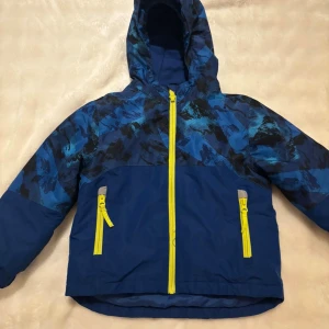 Blå vindjacka från Lupilu med huva/ kurtka  - Blå vindjacka från Lupilu med coolt abstrakt mönster i olika blå toner och svarta detaljer upptill. Jackan har huva, två gula dragkedjefickor och matchande gul dragkedja framtill. Perfekt för blåsiga dagar och har elastiska muddar vid ärmslut. 