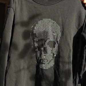 Svart tröja med döskalle från Philipp Plein - Cool svart långärmad tröja från Philipp Plein med glittrande döskallemotiv framtill i små stenar. Det har lossnat 5 pärlor men inget man direkt tänker på. Tröjan har rund hals och en exklusiv metallogo i nacken. Perfekt för dig som gillar edgy och lyxig streetstyle. Nypris 25,590 kr