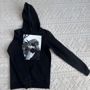 Svart Tupac hoodie med tryck - Svart hoodie med stor svartvit Tupac-tryck på bröstet. Klassisk känguruficka framtill, dragsko i huvan och långa ärmar. 