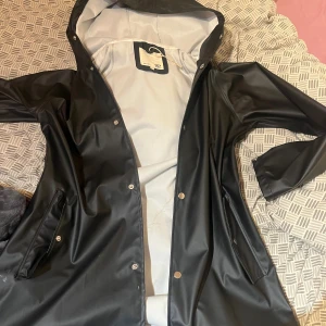 Svart regnjacka med huva från 157 - Svart regnjacka från 157 med huva och tryckknappar framtill. Jackan har två fickor med lock och är helsvart på utsidan med ljus insida. Perfekt för regniga dagar och enkel att matcha med allt. Helt ny 