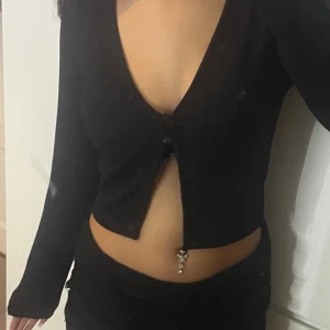 Svart croppad ribbad topp med knappar - Säljer en svart croppad topp med lång ärm och djup v-ringning. Toppen har ribbad struktur och två knappar framtill som ger en öppen detalj vid magen. Perfekt för dig som gillar stilrena och trendiga plagg med lite edge.💕💕pris kan diskuteras