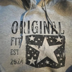 Grå hoodie med stjärntryck och huva - Snygg grå hoodie med tryck framtill där det står 'ORIGINAL FIT EST 2024' och en stor stjärna omgiven av mindre stjärnor. Tröjan har huva med snörning och en klassisk känguruficka. Perfekt för en chill och avslappnad stil.