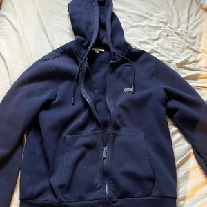 Mörkblå hoodie från Lacoste - Säljer en mörkblå hoodie från Lacoste med dragkedja och klassisk krokodillogga på bröstet. Tröjan har huva med snörning, två stora fickor framtill.