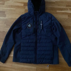 Mörkblå Sail Racing jacka - Snygg mörkblå jacka från Sail Racing med huva och broderad patch på bröstet. Jackan har quiltad framsida och rygg, dragkedja framtill och logga på bröstet. Materialet är polyester och jackan har långa ärmar och en sportig look.