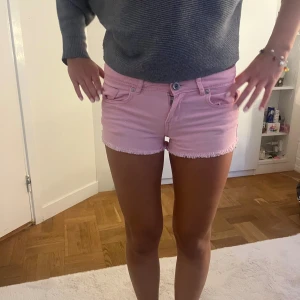 Rosa jeansshorts med fransig kant - Säljer ett par ljusrosa jeansshorts med fransig kant nertill. Shortsen har klassisk femficksdesign, knapp och dragkedja framtill samt bälteshällor. Perfekta för varma dagar och ger en cool vibe till din outfit.