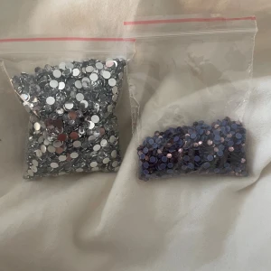 Ansiktsstenar i silver och lila - Två påsar med små ansiktsstenar, en med silvriga och en med lila stenar. Perfekta för att skapa kreativa sminklooks eller festivalmakeup. Stenarna är runda och platta undertill för enkel applicering med lim.