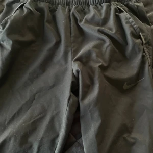 Svarta träningsbyxor från Nike - Svarta träningsbyxor från Nike med elastisk midja och diskret Nike-logga på benet. Byxorna har en enkel och sportig design, perfekta för gymmet eller chill. Tillverkade i ett lätt och bekvämt syntetmaterial.