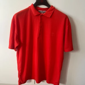 Röd pikétröja från South Lodge strl M/L - Snygg röd pikétröja från South Lodge i storlek M/L. Klassisk krage, knappar framtill och diskret broderad logga på bröstet. Tillverkad i en blandning av polyester och bomull.