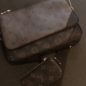 Svart axelväska med monogram - Snygg svart axelväska i skinn med klassiskt monogrammönster. Väskan har flera fack, avtagbar axelrem och matchande mindre plånbok och pouch. Perfekt för dig som vill ha en stilren och lyxig accessoar med ikonisk design.