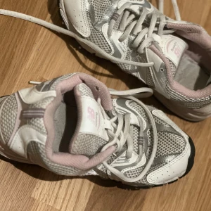 New Balance vita och silver sneakers - Säljer ett par New Balance sneakers i vitt och silver med ljusrosa detaljer. Skorna har meshpaneler för extra andningsförmåga, klassisk snörning och en sportig vibe. Perfekta för dig som gillar retrostil och bekväma skor till vardags.