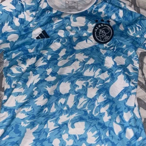 Ajax blåvit fotbollströja Adidas S - Ajax fotbollströja från Adidas i storlek S. Tröjan har ett unikt blått och vitt camouflage-mönster, Ajax-logga på bröstet och Adidas-märke. Tillverkad i lätt och ventilerande material, perfekt för träning eller match.