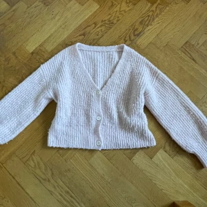 Ljusrosa stickad kofta med knappar - Supermysig ljusrosa stickad kofta med v-ringning och tre vita knappar framtill. Koftan har lång ärm, ribbade muddar och en lite croppad passform. Perfekt att slänga på sig när det är kyligt ute.