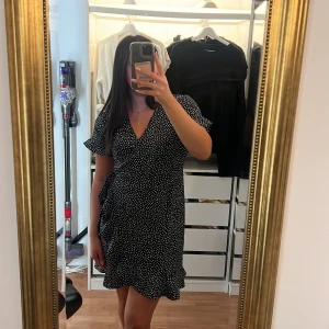 Svart prickig omlottklänning - Supersöt svart kortklänning med vita prickar och omlottmodell. Klänningen har v-ringning, volangdetaljer nertill och korta ärmar. Perfekt för dig som gillar en feminin och trendig stil.