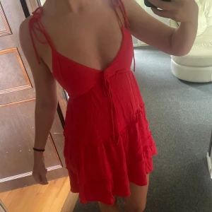 Röd kort sommarklänning med volanger - Supersöt röd kort klänning med volanger och knytband på axlarna. Djup v-ringning och tunna axelband. Perfekt för varma dagar och har en flowy passform som känns både bekväm och trendig. Endast använd 1 gång, nypris 389kr.