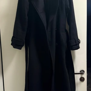 Svart lång kappa från Gina Tricot - Säljer en svart lång kappa från Gina Tricot i storlek XXS. Kappan har bälte i midjan, knappar framtill och snygga detaljer vid ärmsluten. Perfekt för dig som vill ha en klassisk och stilren look under höst och vinter. Passar mig som är S/M. 