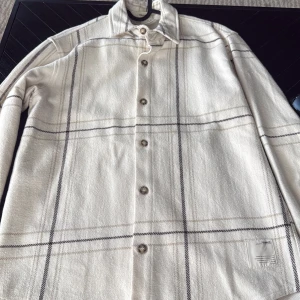 Rutig overshirt från Pull&Bear - Snygg overshirt från Pull&Bear i beige och vitt med rutigt mönster och klassisk krage. Långärmad modell med knappar framtill och mjukt material som känns skönt mot huden. Perfekt att slänga över en t-shirt för en chill look.