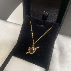 Halsband med svart hjärthänge i guld - Supersnyggt halsband med tunn guldfärgad kedja och ett hjärtformat hänge i svart sten, inramat av guld och dekorerat med en pil genom hjärtat. Perfekt statement-piece för dig som gillar unika smycken med cool vibe. Rostfri kjedjan är 40cm lång 