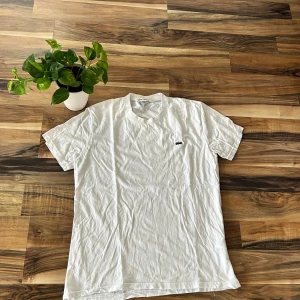 Vit t-shirt från Lacoste, storlek L - Vit klassisk t-shirt från Lacoste i bomull med rund hals och korta ärmar. Ikoniska gröna krokodilloggan broderad på bröstet. Enkel och stilren design som passar perfekt till jeans eller shorts.