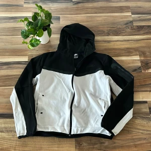 Nike svart tröja  - Nike vindjacka i svart och vitt med huva och hel dragkedja framtill. Jackan har två fickor och diskret Nike-logga på bröstet. Tillverkad i mjukt material som passar perfekt för en sportig och avslappnad stil.