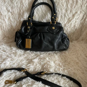 Svart skinnväska Marc by Marc Jacobs - Snygg svart handväska i skinn från Marc by Marc Jacobs med guldfärgade metalldetaljer och avtagbar axelrem. Väskan har ett klassiskt format, dekorativa spännen och en innerfoder med grafiskt mönster. Perfekt för dig som gillar stilrena accessoarer med lyxig känsla.