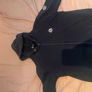 Svart hoodie från Moncler med dragkedja - Säljer en svart hoodie från Moncler med hel dragkedja framtill och huva. Tröjan har Moncler-logga på bröstet och ärmen, samt praktiska fickor på sidorna. Perfekt för dig som gillar stilrena och exklusiva streetwear-plagg. Storlek L men lite för liten för sin storlek.