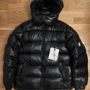 Svart Moncler Maya pufferjacka med huva - Säljer en svart pufferjacka från Moncler med glansig finish och avtagbar huva. Jackan har dragkedja framtill, två fickor med dragkedja och en ärmficka med Moncler-logga. Yttermaterialet är polyamide och fodret består av 90% dun och 10% fjäder. Perfekt för kalla dagar. Använd högst 10 gånger, inga defekter. Märkt med storlek 4, uppskattar till M. Personen på bilderna bär normalt S och är 170 cm. 