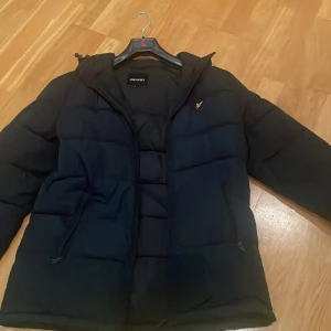 Svart pufferjacka från Lyle & Scott - Svart pufferjacka från Lyle & Scott med huva och dragkedja. Jackan har quiltad design, två fickor med dragkedja och den klassiska Lyle & Scott-loggan i gult på bröstet. Perfekt för kalla dagar och riktigt snygg till streetwear-stilen.