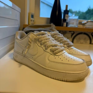 Nike Air Force 1 vita sneakers - Nike Air Force 1 sneakers i helvitt skinn med chunky, tvinnade vita snören. Klassisk låg modell med perforerad tå och rund sula. Perfekta för dig som gillar en clean och tidlös look med en twist på snörningen.