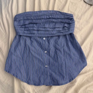 Blåvit randig tubtopp med knappar - Säljer en blå och vit randig tubtopp från Fasster. Toppen är axelbandslös och har knappar framtill samt dragkedja i sidan. Den är sydd i bomullstyg och har en snygg bandeauform med veckad överdel. 