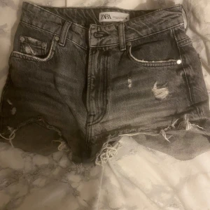 Gråa jeansshorts från Zara - Säljer ett par gråa jeansshorts från Zara med hög midja och råa, fransiga kanter. Shortsen har slitna detaljer och klassisk femficksdesign. Perfekta för en avslappnad och trendig look.