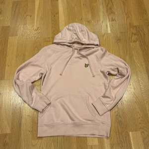 Ljusrosa hoodie från Lyle & Scott - Säljer en ljusrosa hoodie från Lyle & Scott med klassisk gul logga på bröstet. Tröjan har huva med snörning, magficka och långa ärmar. Perfekt för en chill och avslappnad stil.