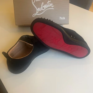 Christian Louboutin svarta sneakers med nitar - Svarta sneakers från Christian Louboutin i mocka med ikoniska röda sulor och svarta nitar på tån. Skorna har snörning och en rund tå. Snygg och edgy design som sticker ut, perfekt för dig som vill ha något extra. Box samt extra nitar tillkommer! Köp innan den 19de september för fri frakt!